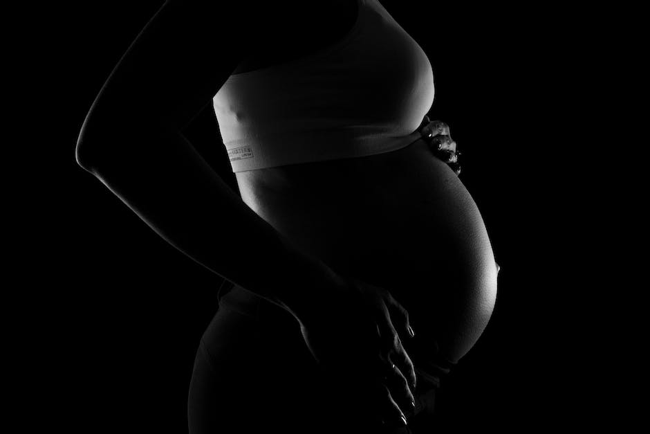 A black and white photograph of a pregnant woman standing at the edge of a river with her hands crossed and her eyes closed. The image has a soft focus and a dreamy quality.